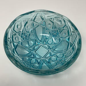 May include: A clear, light blue glass bowl with a detailed, geometric pattern. The bowl's design features diamond, star, and floral shapes. The bowl has a slightly oval shape and is likely vintage.
