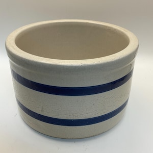 May include: A cream-colored, cylindrical stoneware crock with two horizontal navy blue stripes. The antique container has a wide opening and a slightly textured surface, suggesting age and use. The crock is likely for kitchen or home decor.