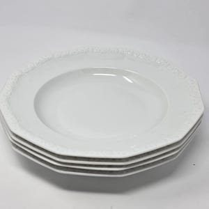 May include: A stack of four white, octagonal-shaped plates. Each plate has a slightly recessed center and a decorative, embossed border. The plates are stacked on top of each other, showcasing their clean, minimalist design.