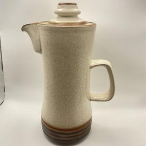 May include: A tall, speckled ceramic coffee pot with a lid and handle. The pot is primarily beige with brown accents on the base and lid. The spout is on the left side. A vintage kitchenware item.