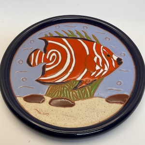 May include: A round ceramic decorative plate featuring a vibrant orange and white fish design. The fish is set against a light blue background with green seaweed and brown rocks. The plate has a dark blue rim.