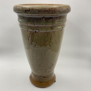 May include: A tall, olive green ceramic vase with a wide rim and a flared base. The vase has a textured surface with visible cracks and imperfections, giving it a rustic appearance. The vase is suitable for displaying flowers or as a decorative accent.