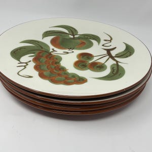 May include: A stack of white ceramic plates with a brown rim. Each plate is decorated with a green and orange fruit design. The fruit includes grapes, apples, and cherries. The plates are stacked on top of each other.