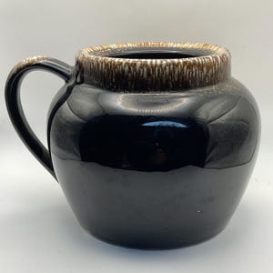 May include: A black ceramic jug with a handle. The rim and handle have a brown and tan glaze. The jug has a glossy finish and a rounded shape. This decorative jug is suitable for serving beverages.