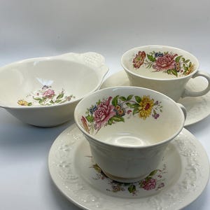 Peut inclure: Un service à thé crème avec un motif floral composé de fleurs roses, jaunes et bleues. Un bol assorti est également présent. L'ensemble a un bord texturé et est posé sur un fond blanc.