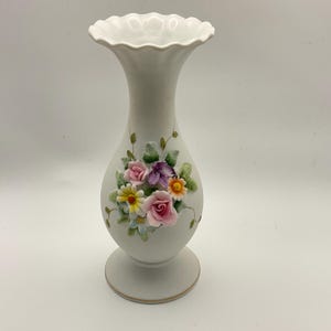 May include: A white porcelain vase with a ruffled rim and a pedestal base. The vase is adorned with a cluster of colorful, three-dimensional flowers, including pink roses, purple petals, and yellow daisies. Delicate green vines and leaves accent the floral arrangement.