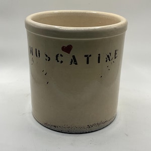May include: A cream-coloured stoneware crock with the word "MUSCATINE" in black lettering and a red heart. The crock has a cylindrical shape with a slightly flared rim and a distressed, vintage appearance.