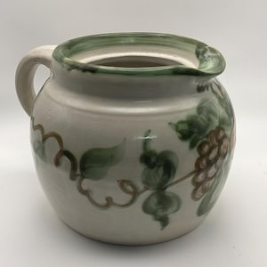 May include: A ceramic pitcher with a handle, painted with green leaves and brown grapes. The rim is painted green, and the pitcher has a cream-colored base. The pitcher is a decorative item.