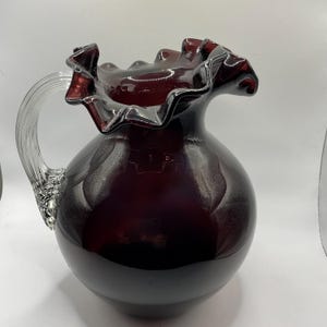 May include: A dark red glass jug with a ruffled rim and a clear, textured handle. The jug has a rounded body and a glossy finish, suitable for serving drinks. The design is elegant and decorative.