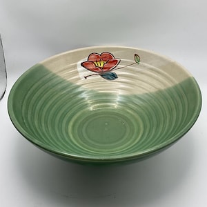 May include: A ceramic bowl with a two-tone design. The top half is beige, and the bottom half is green with concentric circles. A red flower with green leaves is painted on the beige section. The bowl is round and has a wide opening.