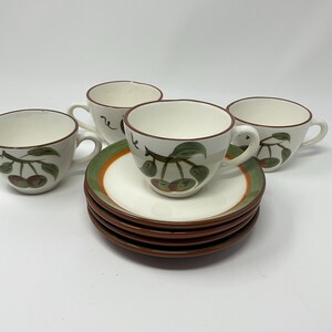May include: A set of four ceramic teacups and saucers. The cups and saucers are white with a brown rim and a cherry design. The saucers have a green and orange border.