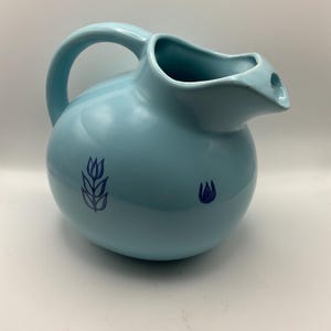 May include: A light blue ceramic pitcher with a rounded body, a curved handle, and a spout. The pitcher features two dark blue floral designs. This decorative pitcher is suitable for serving beverages.
