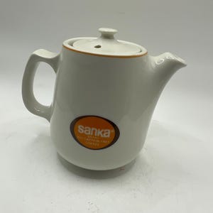 May include: White ceramic teapot with a lid and handle. The teapot has an orange trim and a brown and orange oval logo that reads "sanka" and "97% caffeine free coffee". The teapot is designed for serving coffee.
