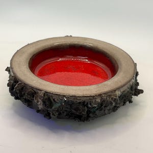 Peut inclure: Un bol en céramique avec un extérieur texturé gris foncé et un intérieur lisse rouge vif. Le bord est de couleur gris clair. Le bol a une forme unique et organique, et est probablement fait à la main.