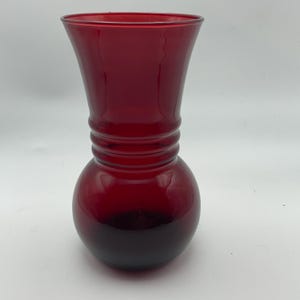 May include: A ruby red glass vase with a bulbous base and a flared top. The vase has a series of horizontal ridges around the middle, adding a decorative element. The glass appears smooth and glossy, reflecting light.