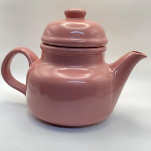 May include: A pink ceramic teapot with a matching lid. The teapot has a rounded body, a curved handle, and a spout for pouring. The lid has a small knob for easy removal. The teapot is a solid, glossy pink color.