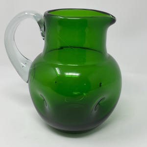 May include: A green glass pitcher with a clear glass handle. The pitcher has a rounded body with a spout for pouring. The handle is attached to the side of the pitcher. The pitcher is made of a glossy glass material.