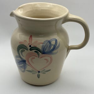 May include: Cream ceramic jug with a curved handle and spout. The jug is decorated with a floral design featuring pink hearts, blue bows, and green leaves. A decorative piece for the kitchen or dining room.