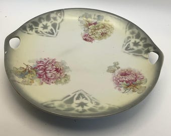 May include: Oval serving tray with a gray rim and handles. The tray features a cream-colored center with pink and yellow floral designs. Gray floral patterns are on the rim. The tray is likely made of ceramic or porcelain.