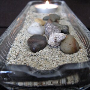 May include: A clear glass tray with a sand base and a lit candle in the center. The tray is decorated with small stones and rocks.