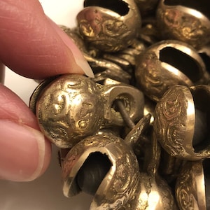 May include: Close-up of a cluster of small, antique-looking bells. The bells are a tarnished gold color with intricate embossed patterns. Each bell has a dark interior and is connected to others by a metal loop.