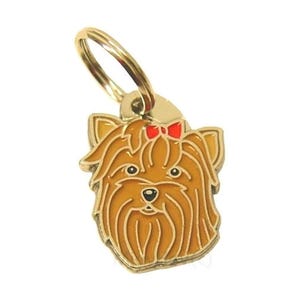 Puede incluir: Una chapa de perro de metal dorado con una imagen de dibujos animados de un Yorkshire Terrier con un lazo rojo. El perro tiene el pelo marr&oacute;n y la nariz negra.