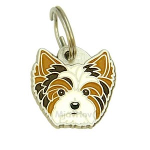 May include: A dog tag featuring a Yorkshire Terrier design. The tag has a silver-colored ring for attachment. The dog's face is depicted with brown, white, and black accents. The text "Mia-Tev" is visible at the bottom.