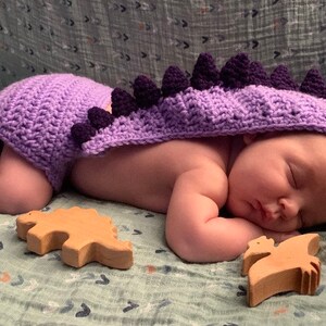 May include: A baby wearing a purple crocheted dinosaur costume with a hood, lying on a green blanket with wooden dinosaur toys.