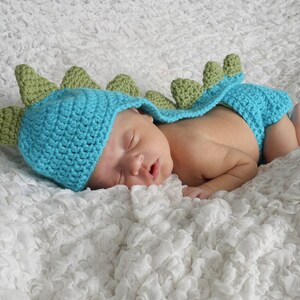 May include: A baby wearing a blue and green crocheted dinosaur costume, sleeping on a white textured blanket.
