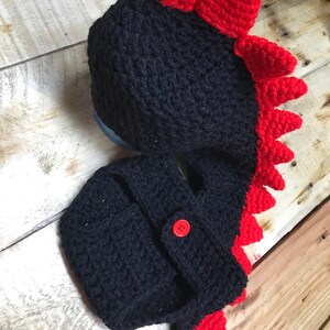May include: Black and red crocheted baby hat and nappy cover with a dinosaur design. The hat has a red spiky crest and the nappy cover has a red tail.