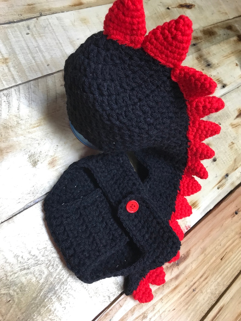 May include: Black and red crocheted baby hat and nappy cover with a dinosaur design. The hat has a red spiky crest and the nappy cover has a red tail.