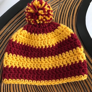 May include: A crocheted hat with a striped pattern in maroon and yellow. The hat has a yellow pom-pom on top.