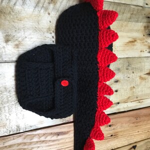 May include: Black crocheted baby dinosaur costume with red spikes and a button fastening.