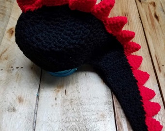 May include: Black crocheted dinosaur hat with a red crocheted spike on top and a long tail.