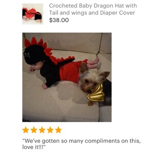 May include: A baby wearing a crocheted red and black dragon costume with a tail and wings. The baby is lying on a sofa with a small dog wearing a gold wing costume.