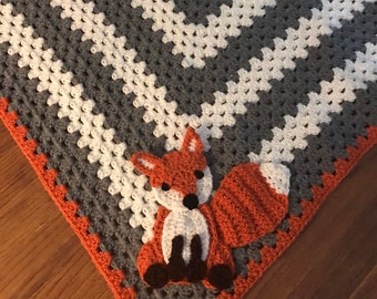 May include: A crocheted baby blanket with a grey and white geometric pattern. A crocheted orange fox is attached to the corner of the blanket.