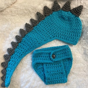 May include: A crocheted dinosaur costume for a baby, featuring a teal blue tail and a matching teal blue nappy with a button. The tail has grey spikes.