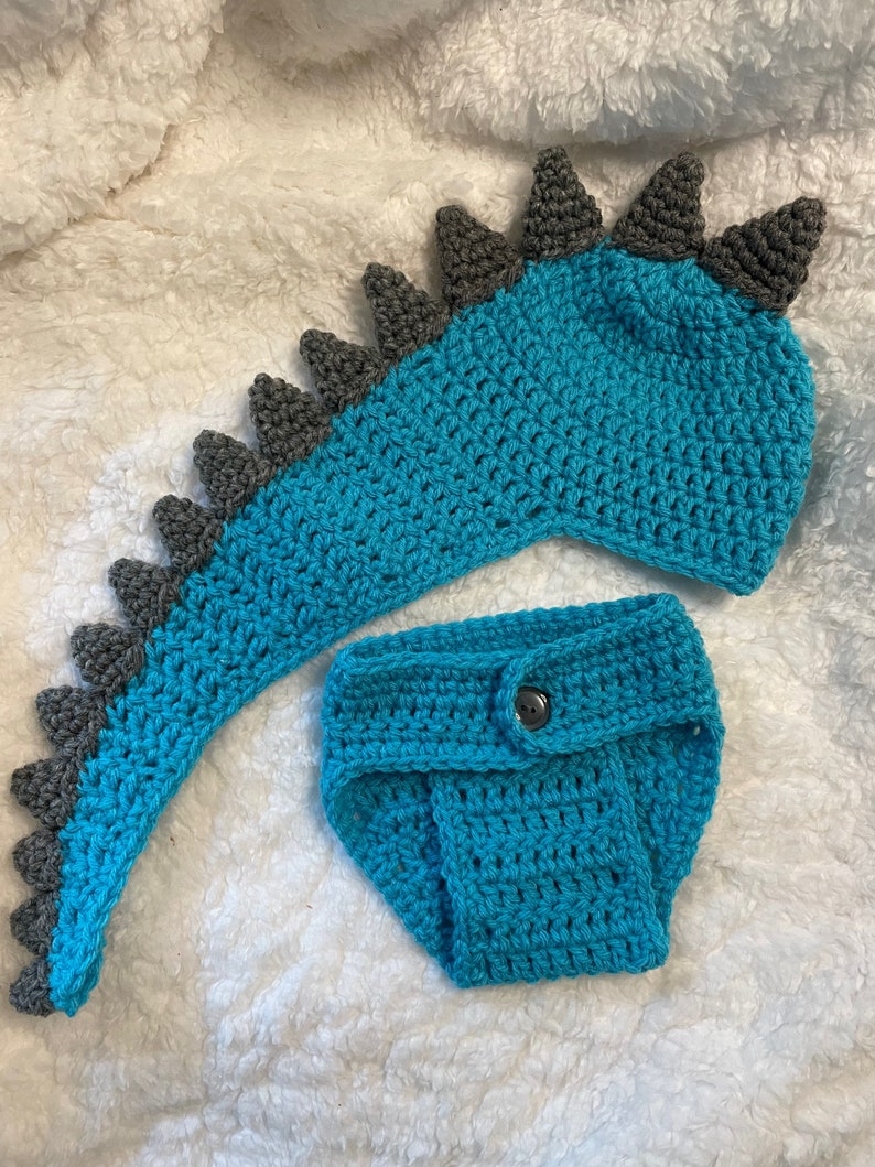 May include: A crocheted dinosaur costume for a baby, featuring a teal blue tail and a matching teal blue nappy with a button. The tail has grey spikes.