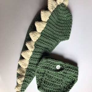 May include: Green and white crocheted dinosaur costume for a baby. The costume includes a hooded hat with a long tail and a matching nappy cover.