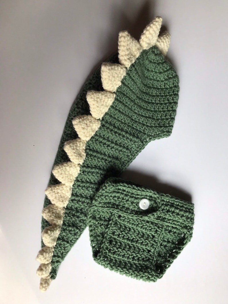 May include: Green and white crocheted dinosaur costume for a baby. The costume includes a hooded hat with a long tail and a matching nappy cover.