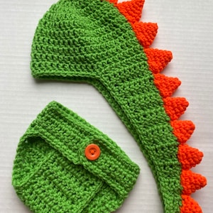 May include: Green and orange crocheted dinosaur hat and nappy set for a baby. The hat has a long tail and spikes on the top. The nappy has a button on the front.