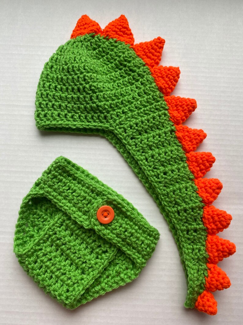 May include: Green and orange crocheted dinosaur hat and nappy set for a baby. The hat has a long tail and spikes on the top. The nappy has a button on the front.