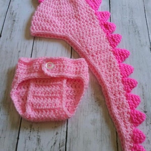 May include: A pink crocheted dinosaur hat and nappy set for a baby. The hat has a long tail and spikes on top. The nappy has a button fastening.