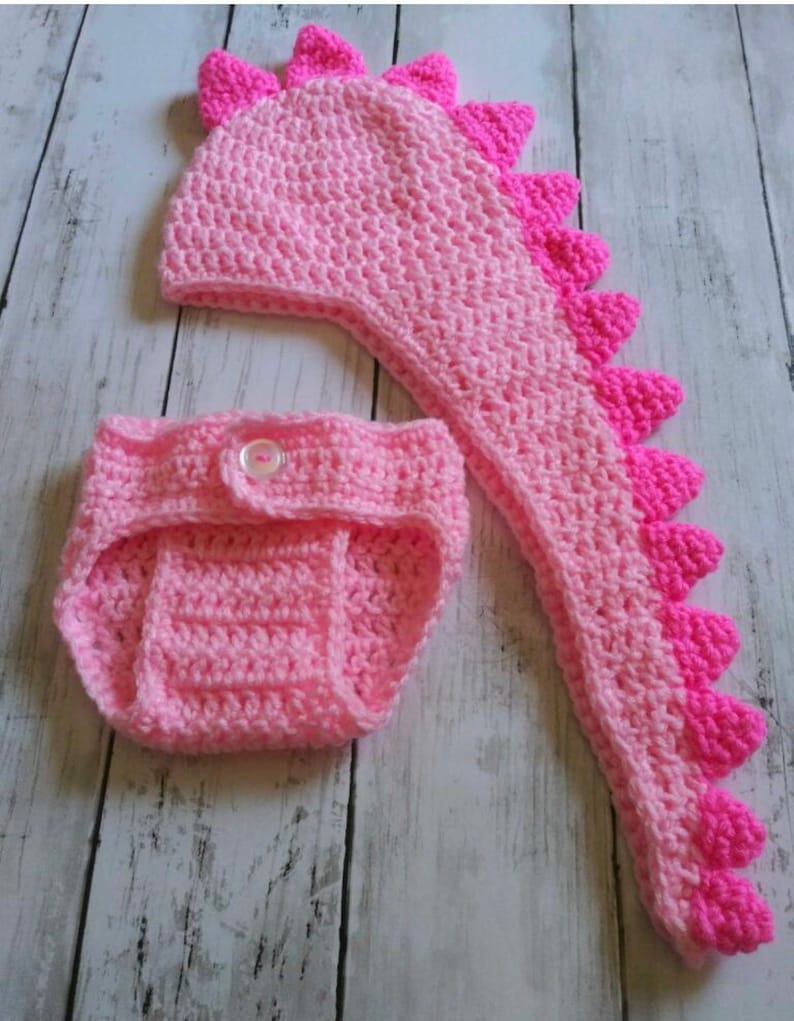 May include: A pink crocheted dinosaur hat and nappy set for a baby. The hat has a long tail and spikes on top. The nappy has a button fastening.