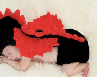 May include: A newborn baby wearing a black and red crocheted dragon costume. The costume features a hat, wings, and a tail.