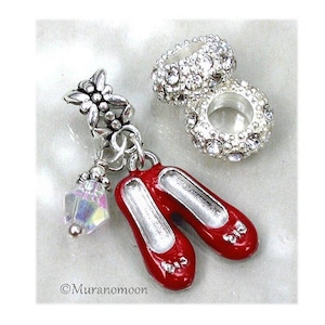 May include: A silver-toned charm featuring a pair of red ballet slippers, a small iridescent crystal bead, and a butterfly-shaped bail, accompanied by two round silver-toned spacer beads encrusted with clear rhinestones. Text reads Muranomoon.