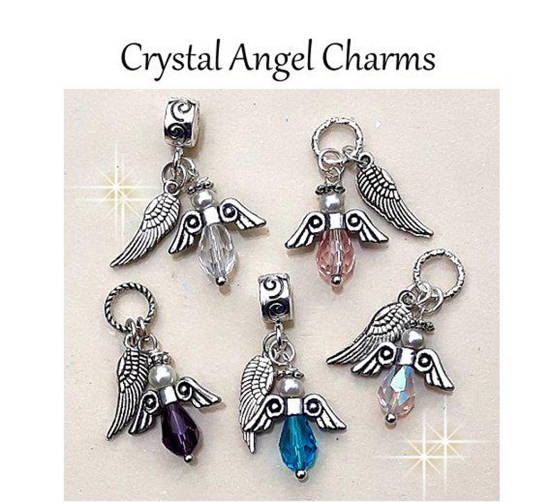 Puede incluir: Crystal Angel Charms. Cinco colgantes de &aacute;ngel plateados con cuentas de cristal en colores transparente, rosa, morado, azul e iridiscente. Cada colgante presenta alas y una cuenta. El texto "Crystal Angel Charms" est&aacute; en la parte superior.
