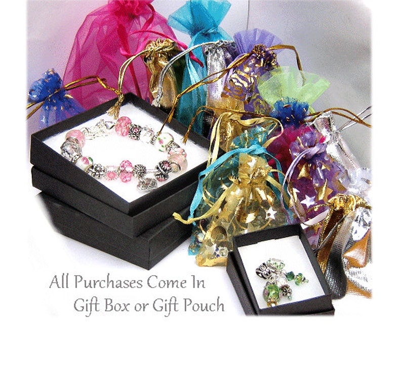 Puede incluir: Un brazalete con cuentas rosas y plateadas exhibido en una caja de regalo negra, rodeado de coloridas bolsas de regalo de organza. El texto "All Purchases Come In Gift Box or Gift Pouch" es visible.