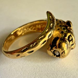 May include: A gold-toned ring featuring a sculpted leopard head design. The band of the ring is textured with dark, recessed spots that mimic the pattern of a leopard's coat, leading to a detailed, three-dimensional leopard head at the front.