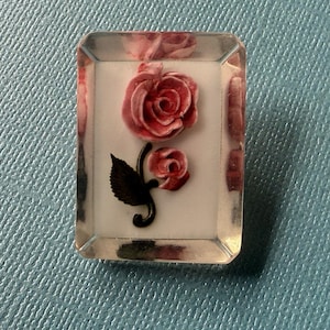 Könnte beinhalten: Rechteckige, transparente Harzbroschen mit einem roten Rosenmuster. Die Brosche zeigt eine große Rose und eine kleinere Knospe auf weißem Hintergrund, mit schwarzen Stielen und Blättern. Die Harzränder sind leicht rosa getönt.